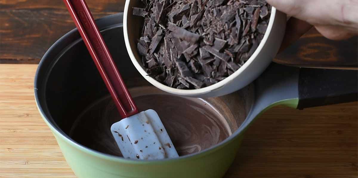 A hand dropping chopped chocolate into a pot with milk and cocoa powder.