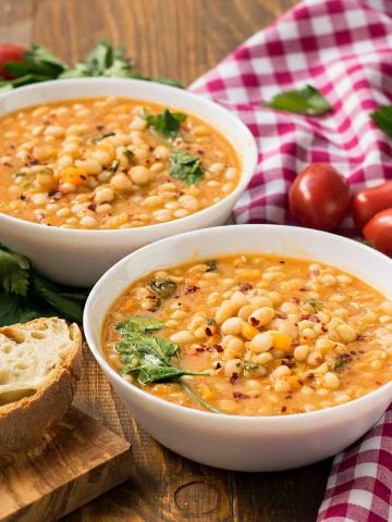 Two bowls of Italian white bean soup, fresh bread on a cutting board and a towel.