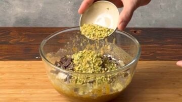 A hand adding ground pistachios to a bowl with cookie dough.