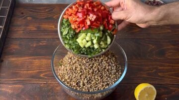 A hand adding chopped vegetables to a bowl with brown lentils.
