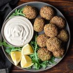 A plate with falafel with a bowl of tahini sauce and lemon wedges.