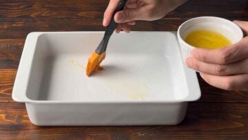 A hand brushes with butter a baking dish.