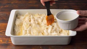 A hand brushes ruffled sheets of phyllo with melted butter.