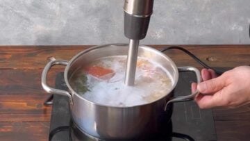 A hand holds an immersion blender and blends the soup.
