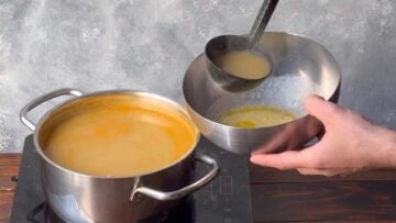 A hand transferring hot soup to a bowl with lemon and egg mixture.