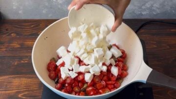 A hand adding tomatoes and feta to the saucepan.