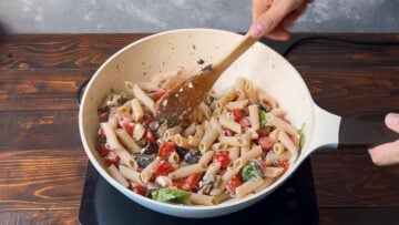 A hand stirring pasta into the saucepan with the eggplant, tomato and feta.