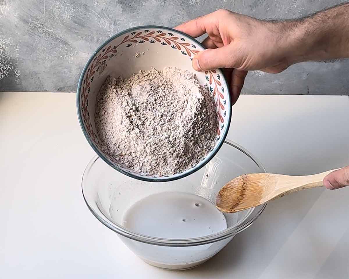 A hand adding whole wheat flour to a bowl with yeast mixture.