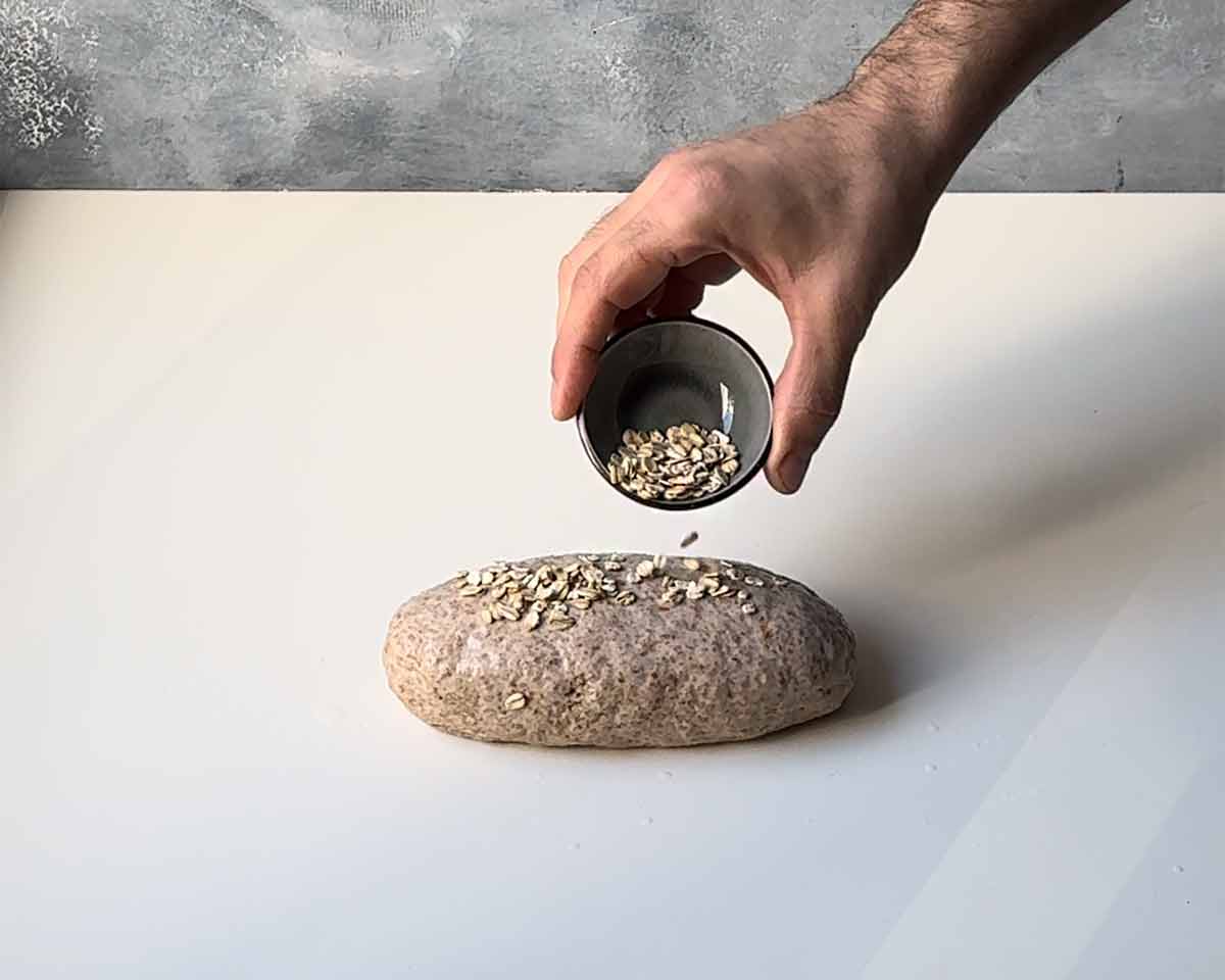 A hand sprinkling oats over the loaf.