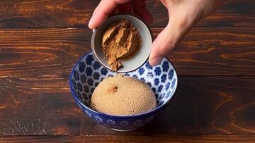 A hand adding cinnamon to a bowl with brown sugar.