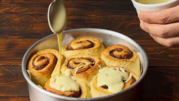 A hand adding white chocolate icing to a pan with cinnamon rolls.