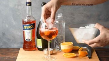 A hand adding ice cubes in a glass with Aperol Spritz.