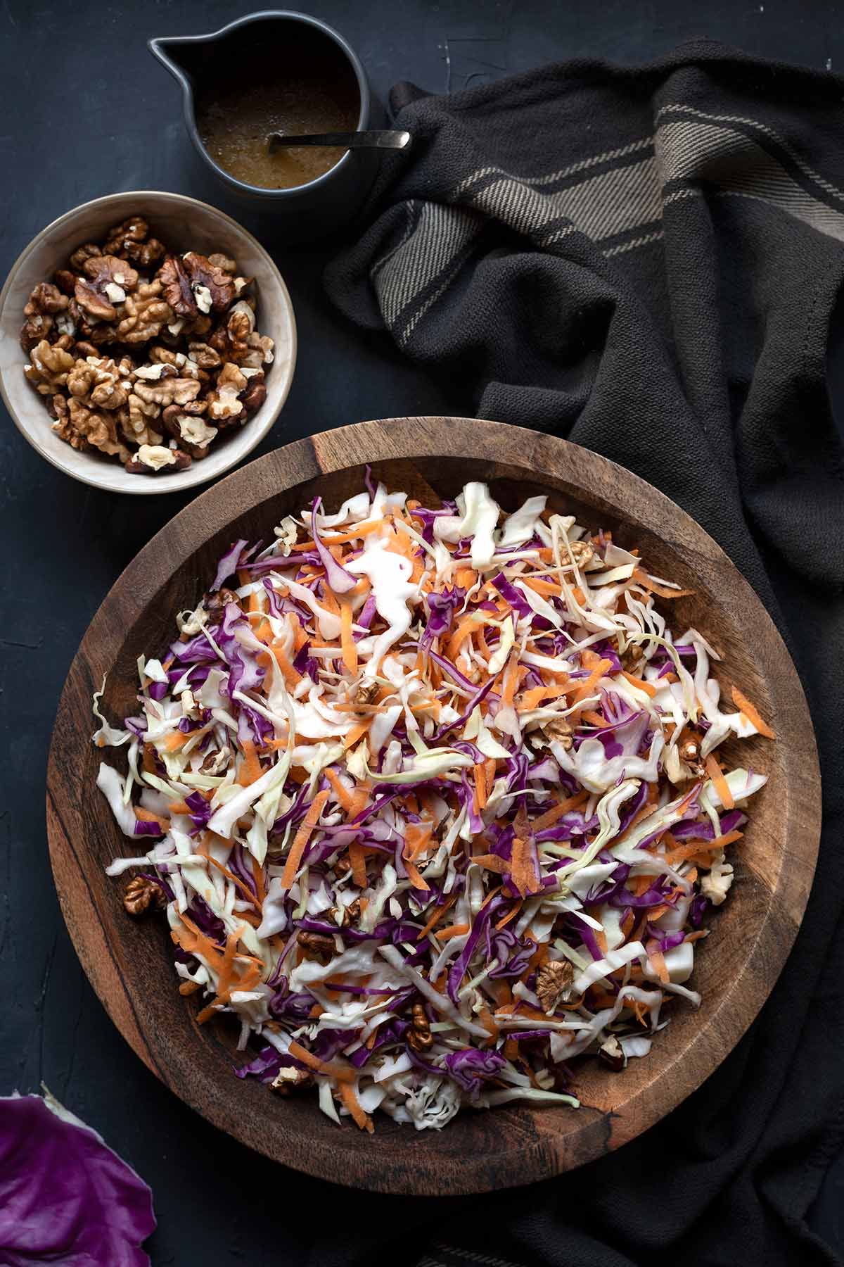 A wooden bowl with Greek cabbage salad, a bowl with balsamic dressing and a bowl with walnuts.