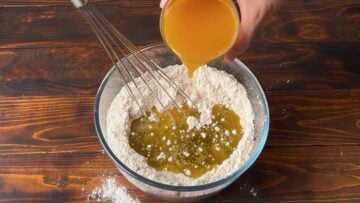 A hand pouring orange juice to a bowl with dry ingredients.
