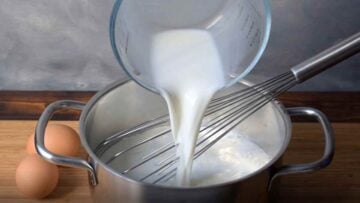 A hand pouring milk in a pot from a jug, a whisk and two eggs.