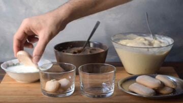 A hand dipping savoiardi biscuits in milk and mont blanc ingredients on the side.