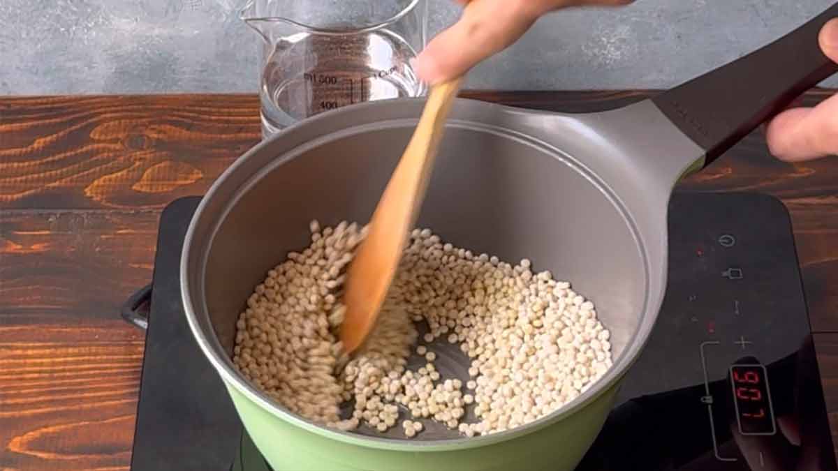 A hand toasting pearl couscous with olive oil inside a pot.