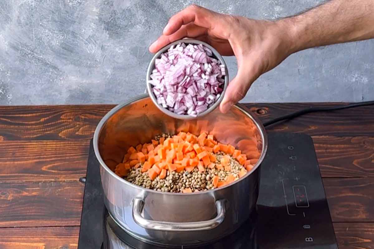 A hand adding chopped onion in a pot with brown lentils and carrot.