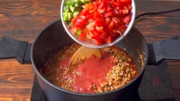 A hand adds chopped bell peppers to the pot with the chili.
