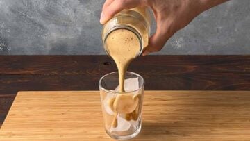 A hand pouring the frothed coffee in a glass with ice cubes.