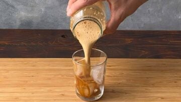 A hand pouring the shaken coffee in a glass with ice cubes.