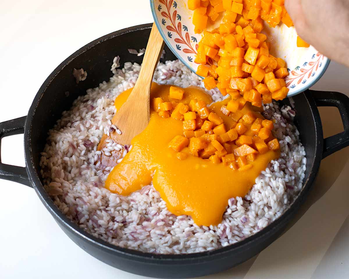 A hand transferring the pumpkin cubes and pumpkin puree into the skillet.