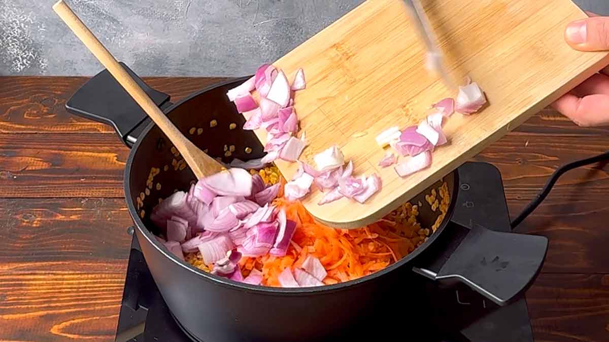 A hand adds chopped onion and grated carrot to the pot.