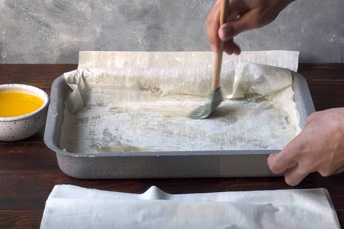 A hand brushing a phyllo sheet with melted butter, inside a baking dish.