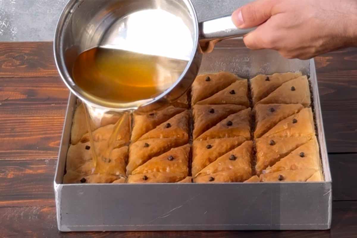 A hand adding hot honey syrup over the hot baklava.