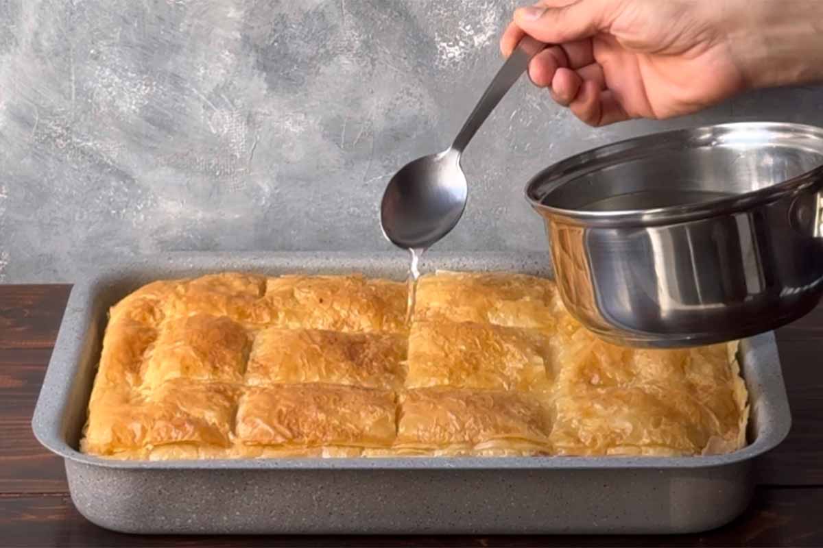 A hand pouring sugar syrup over the hot galaktoboureko.