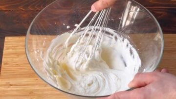 A hand with a whisk beating the butter and sugar mixture in a mixing bowl.