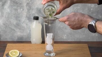 A hand straining the drink over a glass filled with ice cubes.