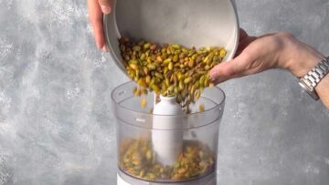 A hand adding peeled pistachios to a food processor.