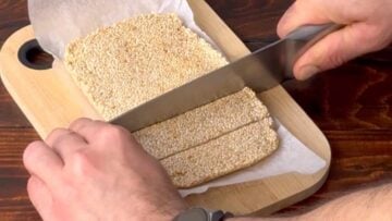 Two hands cutting the sesame mixture into bars with a knife.
