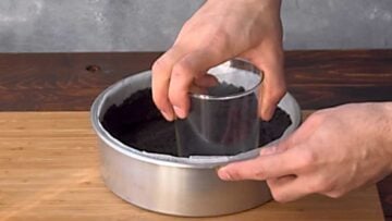 A hand pressing the cookie crumbs with a tumbler into a cake pan.