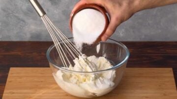A hand adding sugar to a bowl with cream cheese.