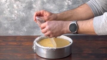 Two hands removing a piece of plastic wrap from the chilled cheesecake before unmolding.