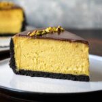 A plate with a slice of pistachio cheesecake with chocolate crust and chocolate topping.