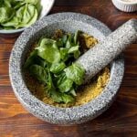 A mortar and a pestle with ground pistachios and basil leaves.