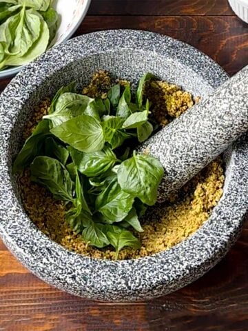 A mortar and a pestle with ground pistachios and basil leaves.
