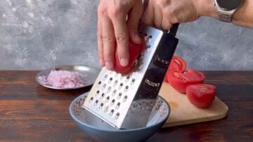 A hand grating a tomato on a box grater.
