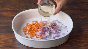 A hand adding wine to a casserole with chopped onion and carrot.