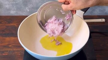 A hand adding grated onion in a saucepan with olive oil.