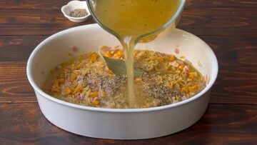 A hand adding chicken broth to a casserole dish with orzo pasta.