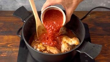 A hand adding pureed tomatoes to a pot with chicken.