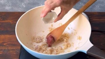 A hand adding tomato paste in a pan with sauteed onion.