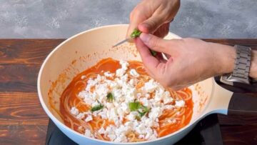 Two hands chopping fresh basil over pasta with tomato sauce and feta.