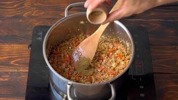 A hand adding black pepper to the pot with bulgur wheat.