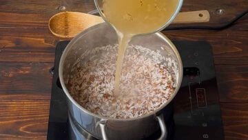 A hand adding broth to a pot with rice and orzo.