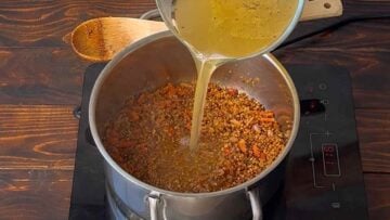 A hand adding broth to a pot with bulgur.